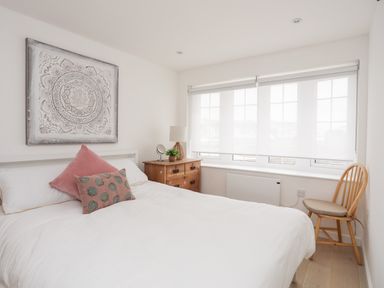 A bedroom with a white bed and pink cushions a wooden chest of drawers with a lamp plant and mirror a wooden chair by a window at 31 Sisial Y Mor in Rhosneigr