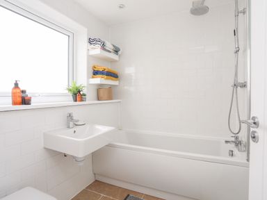 A bathroom with a bathtub shower combination a sink with toiletries folded towels on wall shelves and a window at 31 Sisial Y Mor in Rhosneigr