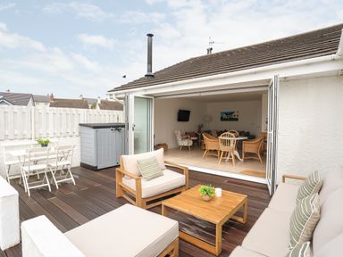An outdoor patio area with wooden furniture and a small white dining set adjacent to an open living room at 31 Sisial Y Mor in Rhosneigr
