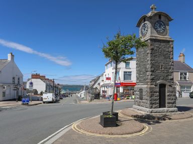 A street with a stone clock tower a small tree and shops near the coast at 31 Sisial Y Mor in Rhosneigr