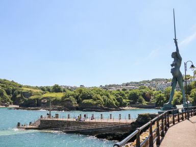 A waterfront scene with a statue holding a sword near a pier and green hills in the background at Mojo in Mullacott near Ilfracombe