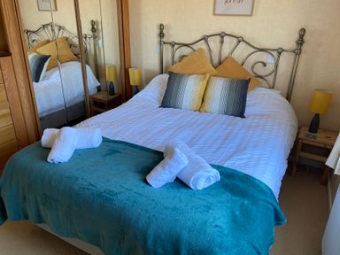 A bedroom with a double bed white bedding striped and yellow pillows blue blanket rolled towels wooden bedside tables with lamps and mirrored wardrobe at Mojo Mullacott near Ilfracombe