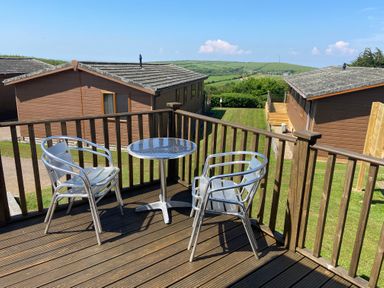 A wooden deck with metal chairs and a table overlooking grassy area and brown wooden cabins at Mojo in Mullacott near Ilfracombe