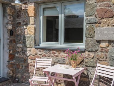 A pink table and two chairs outside a stone cottage with a window and flowers on the table at Traeth Ora in Moelfre