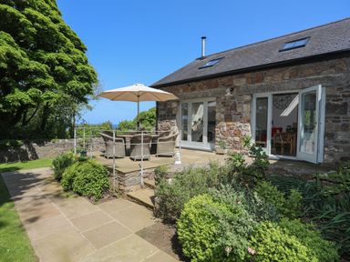 A stone house with an outdoor patio table and chairs under an umbrella surrounded by plants and trees at Traeth Ora in Moelfre