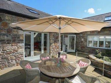 An outdoor patio area with a round wooden table and wicker chairs under a large umbrella at Traeth Ora in Moelfre