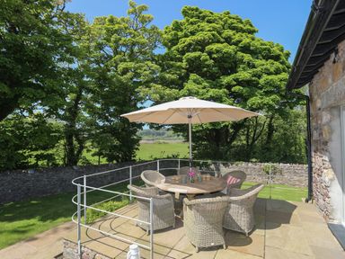 An outdoor patio with a round wooden table surrounded by six wicker chairs and a large umbrella at Traeth Ora Moelfre