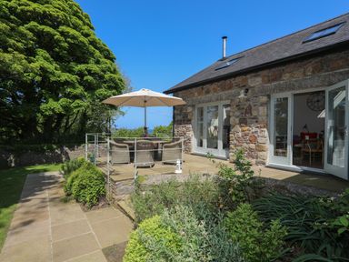 A stone house with a patio table and umbrella surrounded by a garden and trees at Traeth Ora in Moelfre