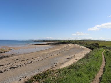 A sandy beach with sea and grassy path alongside at Traeth Ora in Moelfre