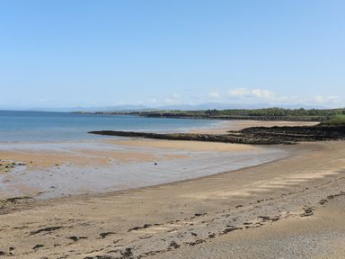 A sandy beach with stretches of water and rocky outcrops with distant hills and blue sky at Traeth Ora in Moelfre
