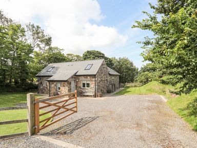 A stone cottage with a pitched roof surrounded by trees and a gravel driveway at Traeth Ora in Moelfre