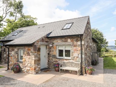 A stone cottage with a bench and flower pots outside on a paved area at Traeth Ora in Moelfre