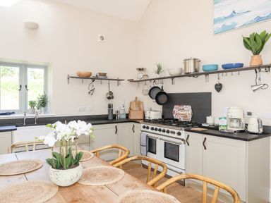 A kitchen with a wooden dining table and chairs a gas stove cabinets and shelves with kitchenware and plants at Traeth Ora in Moelfre