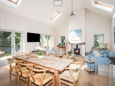 A dining table with six chairs and a flower pot in a room with armchairs a blue sofa a fireplace and skylights at Traeth Ora in Moelfre