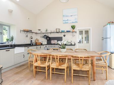 A kitchen with a wooden dining table and chairs a stove and refrigerator and shelves with kitchenware at Traeth Ora in Moelfre