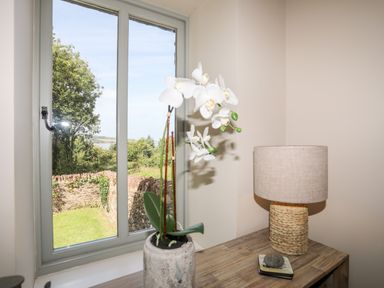 A window overlooking a garden with a stone wall and trees next to a table with a lamp and a potted orchid at Traeth Ora in Moelfre