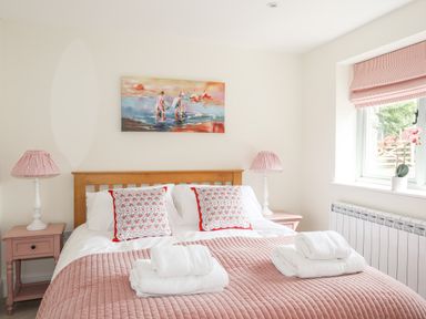 A bedroom with a wooden bed frame pink bedding and pillows two bedside tables with lamps and a window with a plant on the sill at Traeth Ora in Moelfre