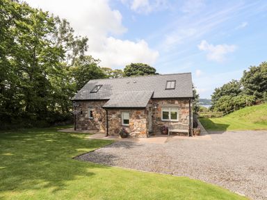 A stone house with a grey roof and two skylights surrounded by green grass and trees at Traeth Ora in Moelfre