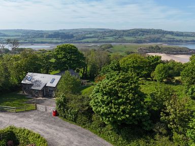 A stone house surrounded by trees near a grassy area and a beach with hills in the background at Traeth Ora in Moelfre