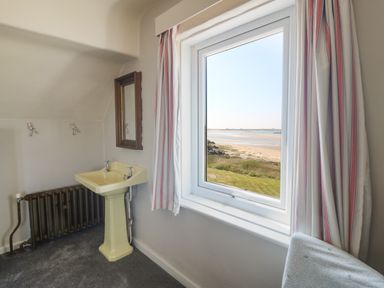 A room with a yellow sink, brown radiator, mirror, window with striped curtains overlooking a beach at Wig Carna in Holyhead