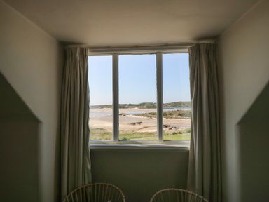 A window with curtains overlooking a sandy beach and vegetation at Wig Carna in Holyhead