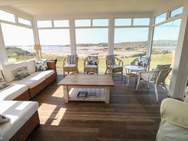 A sunroom with wicker furniture and a wooden coffee table overlooking a beach and grassy landscape at Wig Carna in Holyhead