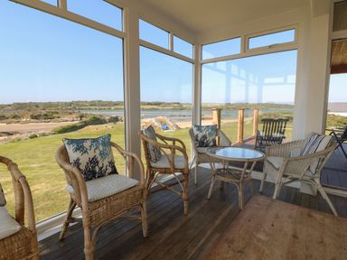A sunroom with wicker chairs and a glass table overlooking a garden and water at Wig Carna in Holyhead
