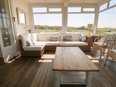 A sunroom with a wicker sectional sofa with cushions and pillows a wooden coffee table two wicker chairs and large windows showing an outdoor grassy landscape at Wig Carna in Holyhead