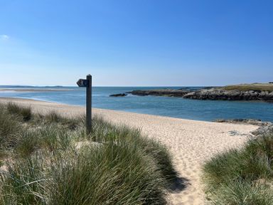 A sandy beach with a walking path sign and grassy dunes by the sea at Wig Carna in Holyhead