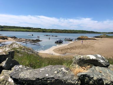 A sandy beach with boats on the water and people walking near the shore at Wig Carna in Holyhead