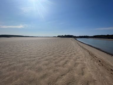 A sandy beach with ripples and a narrow body of water on the right under a clear blue sky at Wig Carna in Holyhead