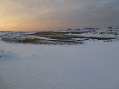 A snow-covered landscape with a partially frozen body of water under a cloudy sky at Wig Carna in Holyhead