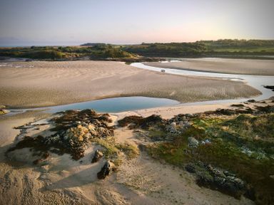 A sandy beach with rocky areas and a tidal pool with green vegetation and hills in the background at Wig Carna in Holyhead