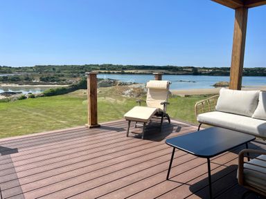 An outdoor deck with lounge chairs and a table overlooking a grassy area and water at Wig Carna in Holyhead