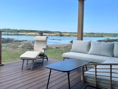 A wooden deck with a lounge chair a sofa and a table overlooking a body of water and grassy landscape at Wig Carna in Holyhead