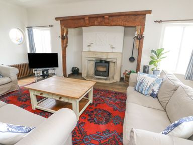 A living room with beige sofas and blue patterned cushions a wooden coffee table a red patterned rug a fireplace with a wood stove a television and windows at Wig Carna in Holyhead
