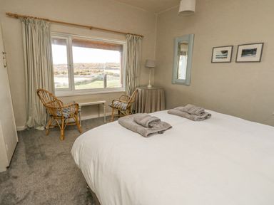 A bedroom with a double bed and two towels on it two wicker chairs by a window with curtains a small table a round side table with a lamp and a mirror on the wall at Wig Carna in Holyhead