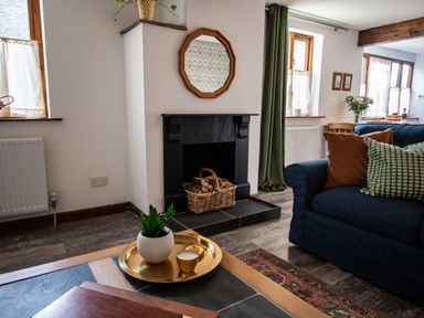 A living room with a black fireplace and a basket of firewood a blue sofa with pillows and a coffee table with a plant and candle at Fern Cottage Muddiford near West Down