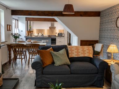 A living area with a dark sofa with cushions and a blanket a wooden dining table and chairs and a kitchen in the background at Fern Cottage in Muddiford near West Down