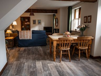 A dining area with a wooden table and chairs next to a living room with sofas and a TV at Fern Cottage in Muddiford near West Down