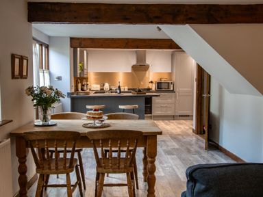 A kitchen and dining area with a wooden table and chairs a cake on the table and kitchen island with stools at Fern Cottage in Muddiford near West Down