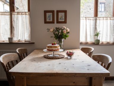 A wooden dining table with a cake and bowl of strawberries with four chairs and flowers on the table at Fern Cottage in Muddiford near West Down