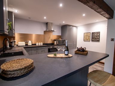 A kitchen with grey cabinets a black countertop appliances and bar stools at Fern Cottage in Muddiford near West Down