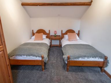 A bedroom with two single wooden beds with gray throws and peach pillows with a wooden nightstand and lamp between at Fern Cottage Muddiford near West Down