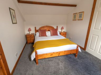 A bedroom with a wooden bed with pillows and a yellow throw and two wooden nightstands with lamps at Fern Cottage in Muddiford near West Down