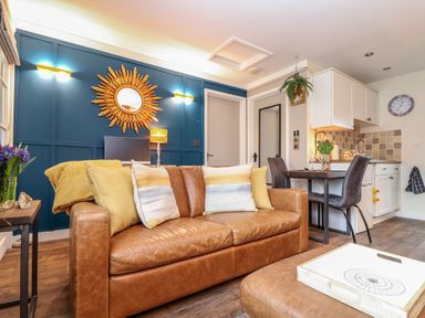 A living room with a brown leather sofa and yellow cushions next to a small dining area and white kitchen cabinets at Inny Brook Cottage in Davidstow near Camelford