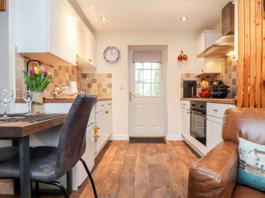 A kitchen with white cabinets a brown tiled backsplash a wooden table with chairs and a brown sofa at Inny Brook Cottage in Davidstow near Camelford