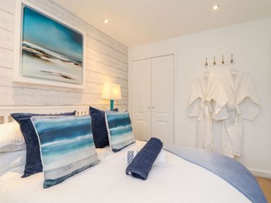 A bedroom with a bed featuring blue pillows and towels a lamp on a bedside table robes hanging on hooks and a painting on the wall at Inny Brook Cottage in Davidstow near Camelford