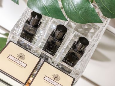 Three small bottles labeled Noir Amber Mandarin and Sandalwood and two boxes labeled Vanity Kit and Shower Cap in a clear organizer with green leaves in the background