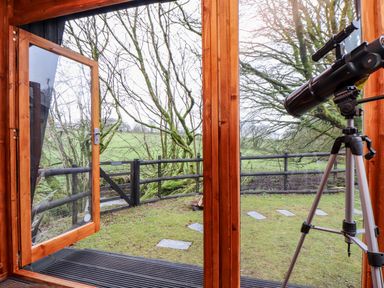 A wooden framed open door leading to a small garden with grass and stone paths a black telescope on a tripod is inside looking outside at Inny Brook Cottage in Davidstow near Camelford